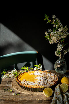 Freshly baked homemade lemon pie with floral crust decorations  in baking form, lemons and cherry blossom  on old wooden table on dark background. 