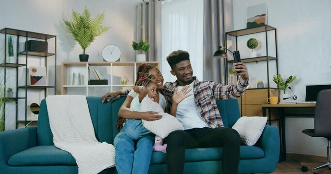 Handsome african man taking selfie on modern smartphone with his beautiful wife and pretty daughter. Young family sitting together on couch, waving hands, hugging and smiling.
