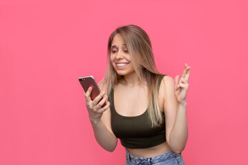 A young pretty caucasian smiling blonde woman crosses her fingers for good luck looking at the phone screen waiting for the results of the lottery or exams isolated on a bright color pink background