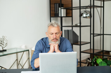 Contemporary senior grey hair businessman sits in front of the laptop contemplating and researching in the office, looking for new ideas or tasks solving, focused mature man in smart casual shirt