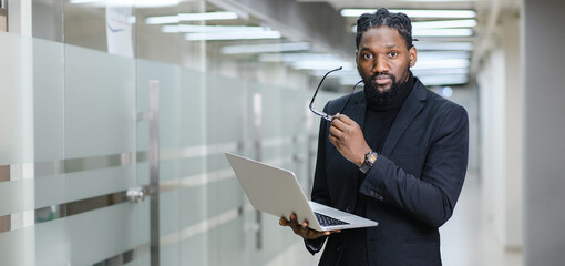 Close-up of a dreamy African-American man with glasses, considering new opportunities or an online project strategy, standing with a laptop against the background of a modern office
