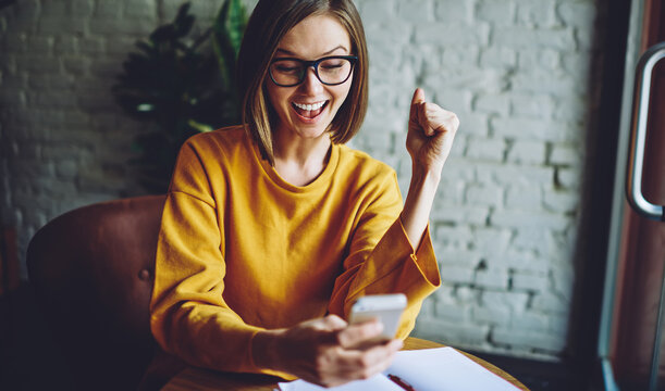 Cheerful Female In Classic Eyeglasses Happy With Received Email Message Using Cafe Internet Connection On Cellular Technology, Smiling Blogger Reading Online Reviews Browsed On Smartphone Device