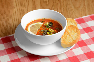 Solyanka, Russian soup with meat, olives and sour cream. served in a white bowl over red plaid tablecloth.