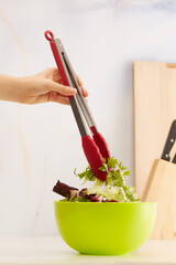 Subject shot of female hand holding stainless steel kitchen tongs with salad leaves above green bowl. Photo is made in kitchen interior. 