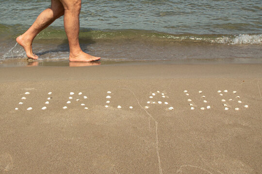 Beine mit Füßen am Meer auf Sand gehen, am Meer, an der Ostsee mit Wellen und Aufschrift "Urlaub" aus Muscheln als Hintergrund
