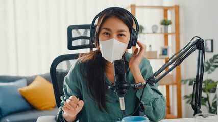 Teenager Asia girl record podcast use headphones and microphone wear mask protect virus look at camera talk in her room. Female podcaster make audio podcast from her home studio, Stay at home concept.