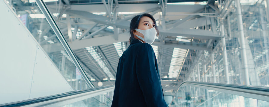 Asian business girl wear face mask drag luggage stand on escalator look around walk to terminal at international airport. Business travel social distancing concept. Panoramic banner background.