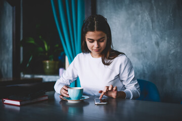 Fototapeta premium Young woman browsing smartphone and having hot beverage in cafeteria