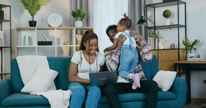Afro american parents playing with little daughter during free time at home. Young couple sitting on couch and using modern laptop for entertaining.