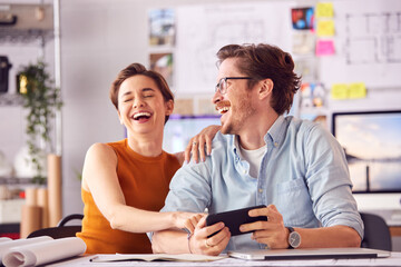Male And Female Architects In Office Sitting At Desk Laughing At Selfie On Mobile Phone