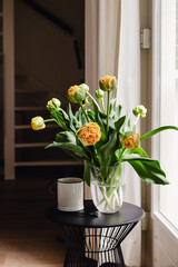 A bouquet of fresh orange tulips and a white mug in a transparent vase on a coffee table next to a window with curtains in a modern style apartment interior. Home decor for slow life.