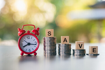 A red alarm clock and a wooden block on a pile of coins that read the word SAVE, concept, time and money saving.