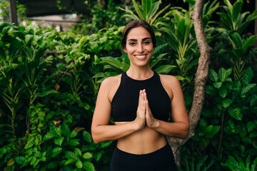 Half length portrait of cheerful Caucasian female yogi in namaste pose smiling at camera during...