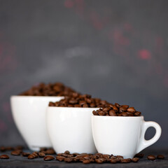 Roasted Coffee beans in white cups different sizes, from smallest to largest, on grey blurred background with copy space at top of image. Close up shot, side view. Soft focus.