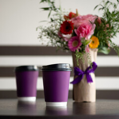 Coffee drink in paper cups on table. Purple disposable cardboard cups and bouquet of roses on blurred background. Coffee to go concept. Soft focus. Square format with copy space. Blank paper cup.
