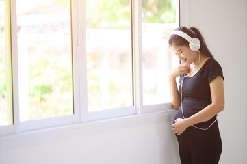 Obraz na płótnie Canvas A pregnant woman in a black dress, wearing a white headphones, stood listening to music by the window.A pregnant woman standing by the window listening to nostalgic music