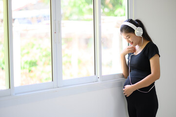A pregnant woman in a black dress, wearing a white headphones, stood listening to music by the window.A pregnant woman standing by the window listening to nostalgic music