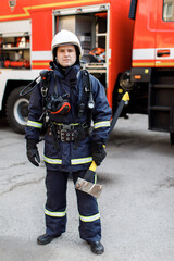 Portrait of serious and confident caucasian fireman standing and holding hammer, wearing special protective uniform in the truck background