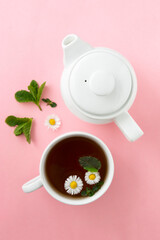 Cup and teapot of herbal tea with chamomile flowers and mint leaves on pink background