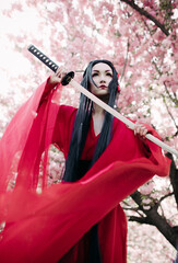 Young woman portrays geisha with sword in her hand near blooming sakura trees.