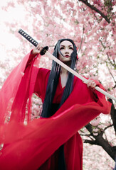 Young woman portrays geisha with sword in her hand near blooming sakura trees.