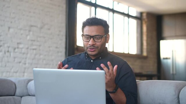 Mixed-race indian man in casual shirt sits on the couch in cozy apartment and using laptop for video connect, talking online with colleagues or family, holding video call on the computer