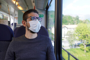 Fototapeta na wymiar Travel safely on public transport. Young man wearing surgical mask looking through train window. Train passenger with protective mask looking through the window.