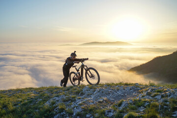 Obraz premium Mountain Biker Pushing Bike Up to the Mountain Above the Clouds at Sunset.