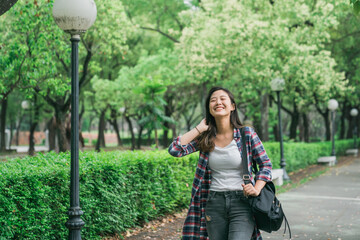 Attractive young asian korean woman enjoying time outside in park with sunshine on face. charming girl with bag walking on pathway cheerful laughing and flicks hair. people breathing fresh air
