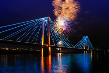 Festive fireworks in the night sky above the illuminated bridge with colorful water reflection. Fireworks in honor of Victory Day in Krasnoyarsk, Russia