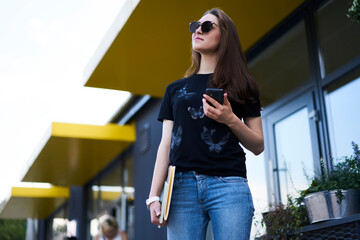 Contemplative female dressed in casual wear thoughtful looking away and thinking about education information, pensive hipster girl in sunglasses holding modern cellphone device and sketch- textbook