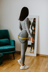 Fitness Woman Measuring butt With Tape at home. strong buttocks concept. Beautiful woman checking success of weight loss program and measuring butt and hips in front of mirror in living room. 