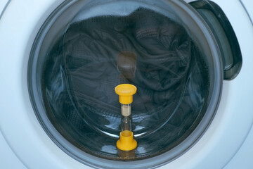 Yellow sandglass on window of washing machine with grey bedspread inside is working, laundry at home. Modern technologies in everyday life, housework and household.