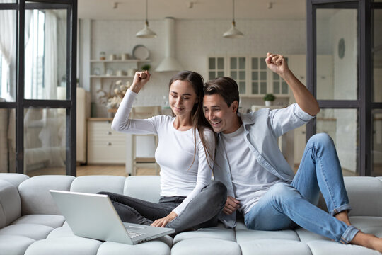 Happy Overjoyed Couple Celebrating Online Betting Bid Lottery Win Sitting On Couch At Home With Raised Hands.