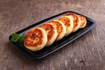 Cottage cheese pancakes or syrniki on black plate decorated with mint, closeup view. Russian, Ukrainian cuisine. Healthy tasty breakfast. Old wood background.