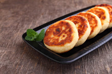 Cottage cheese pancakes or syrniki on black plate decorated with mint, closeup view. Russian, Ukrainian cuisine. Healthy tasty breakfast. Old wood background.