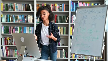Distance training, online lecture. Young smart african american female teacher conducts online lesson for students via video conference uses computer, stand at library, shows information on whiteboard