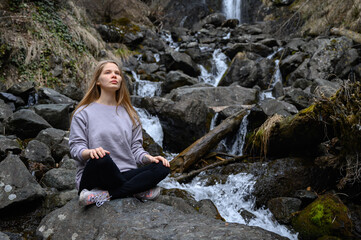 Naklejka na ściany i meble Girl sits on a large stone and meditates near a waterfall next to a mountain river.