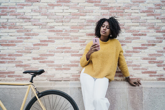 Young stylish black woman using smartphone and social media next to a modern yellow bike in front of a pink brick wall - Powered by Adobe