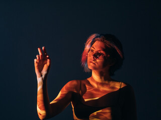 Spiritual enlightenment. Art portrait. Inner peace. Esoteric aura. Curious relaxed woman in eyeglasses reaching out hand in red orange projector light on skin isolated on dark blue.