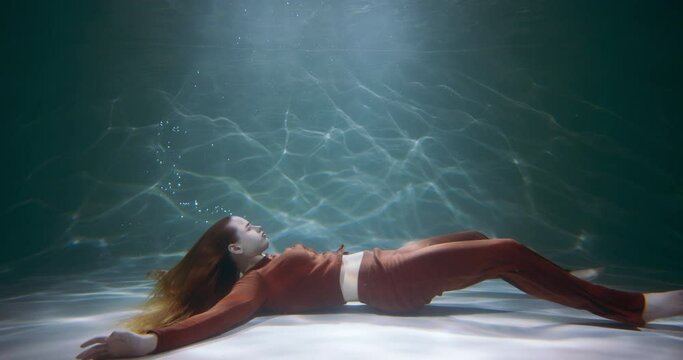 Waking up from sleep. Young beautiful woman lying down underwater on pool floor rises up to get some air slow motion.