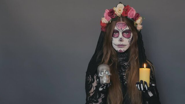 woman in the image of death Santa Muerte or sugar mexican skull with a burning candle and a skull in her hands on a gray background. halloween make-up