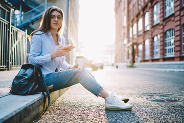 Portrait of attractive brunette woman with takeaway caffeine beverage recreating in city holding mobile phone, young female student with backpack looking at camera during coffee break outdoors