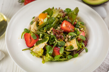 Salad with strawberries, brie cheese, tomatoes and greens. On a white plate and white table.
