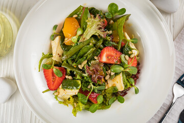 Salad with strawberries, brie cheese, tomatoes and greens. On a white plate and white table.
