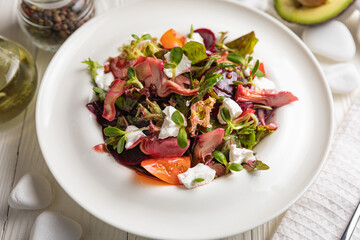 Salad of veal, goat cheese, greens. On a white plate and white table.
