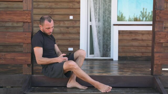 A man in shorts and a T shirt scroll the Internet in the phone sitting on the terrace at the house A businessman in a sports uniform writes a message in the phone sitting on the porch Self isolation