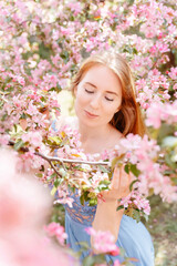 Fototapeta premium Portrait of a cute red-haired girl near a blossoming tree with pink flowers in the garden. Apple blossoming park in spring.