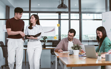 Asian handsome business man and beautiful woman wearing casual shirt, discussing marketing creative idea and walking in hallway of indoor office while colleagues working and meeting at desk.
