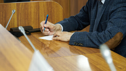 An elderly man - politician, lawyer or teacher writes on a piece of paper while sitting at a table in the boardroom. A court hearing or meeting of politicians and officials